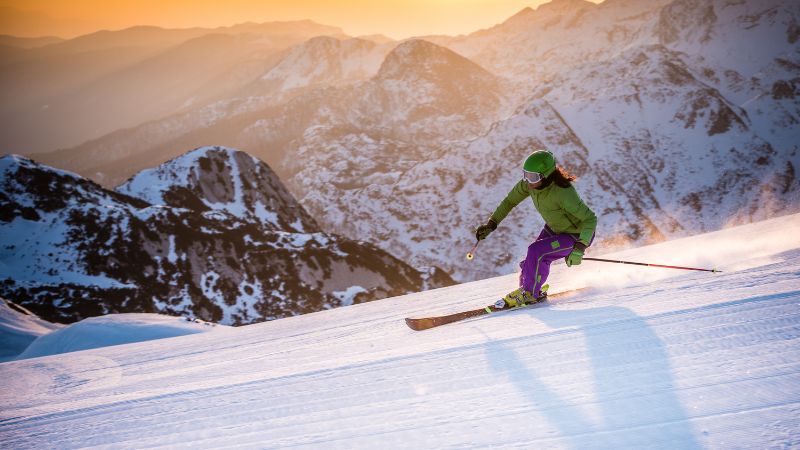 Skier in green and purple gear skiing down a snowy mountain at sunset.