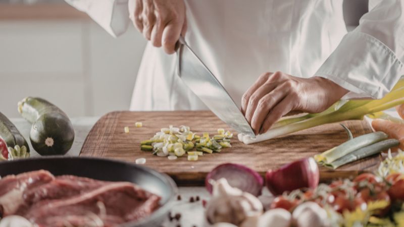 Chef slicing green onions on a cutting board surrounded by fresh vegetables and raw meat.