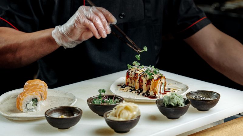 Chef in black uniform plating sushi rolls with microgreens and sauces on a white counter.