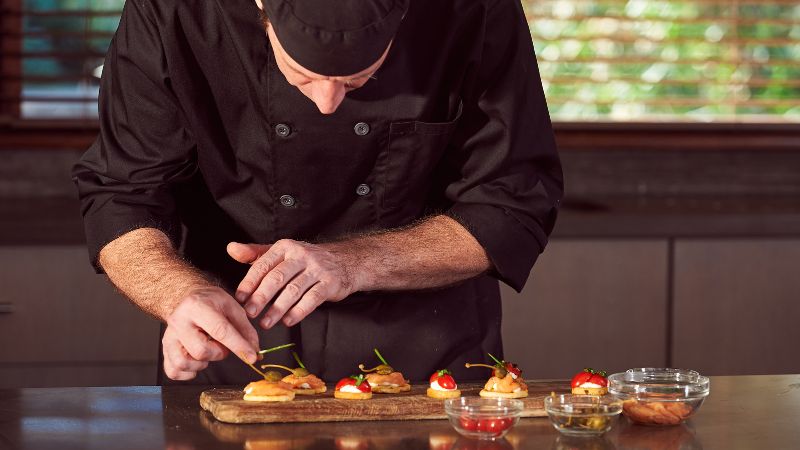 Chef carefully garnishing small appetizers on a wooden board in a kitchen.