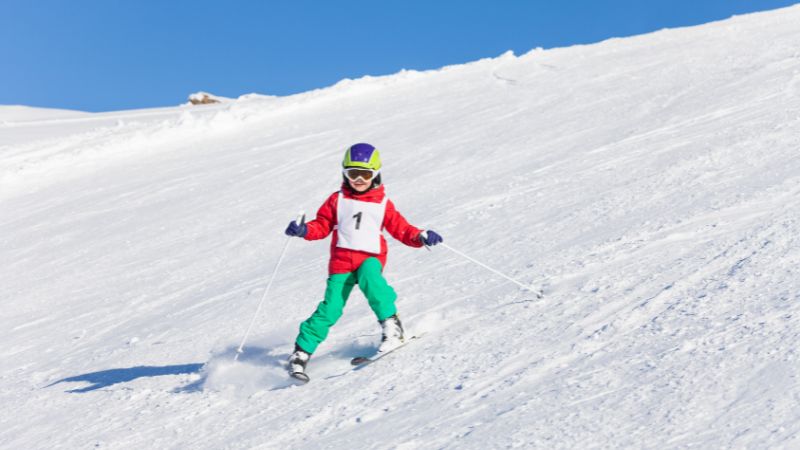 A young boy skiing in the mountains with a safety helmet, goggles, and poles.