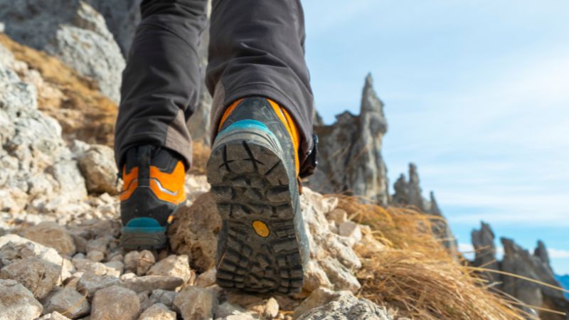 A close-up view of a hiker's feet wearing sturdy hiking boots on a rocky mountain trail. The focus is on the boots and the rough terrain, with small stones and dry grass at the hiker's feet. 
