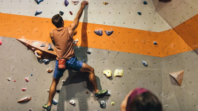 An indoor rock climber is in mid-action, reaching out for the next hold on a climbing wall. The wall features a variety of colorful holds and footholds against a grey surface with an orange stripe pattern. 