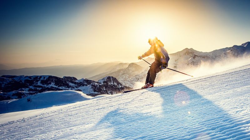 A skier descending a groomed slope at sunset, with dynamic skiing posture and snow spray behind.