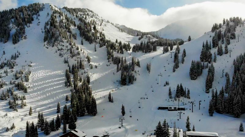 Aerial view of a ski area on a cloudy day with sunlight breaking through, showcasing groomed ski runs, chairlifts, and a base area with buildings. The mountain slopes are dotted with evergreen trees and there are several skiers visible on the slopes.