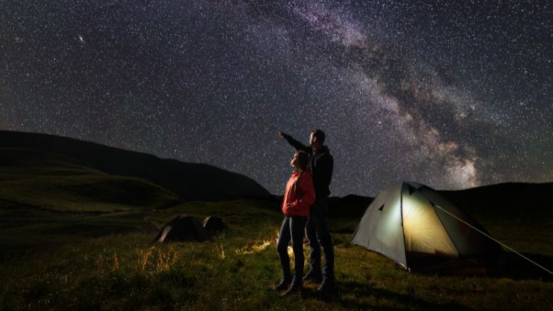 A couple standing beside a tent under a star-filled night sky, with the man pointing toward the stars.