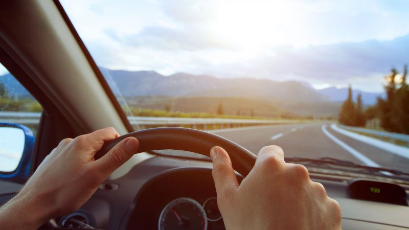 A person drives along a scenic highway with mountains visible in the distance under a bright sky.