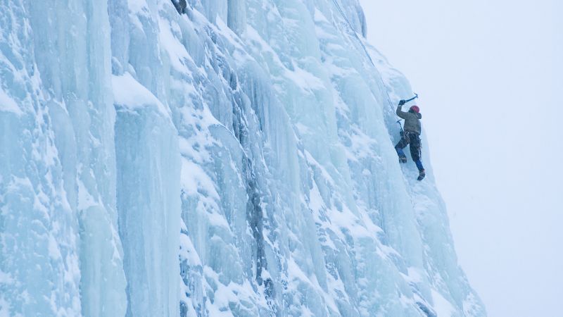 Person ice climbing on a steep, frozen wall using an ice axe.